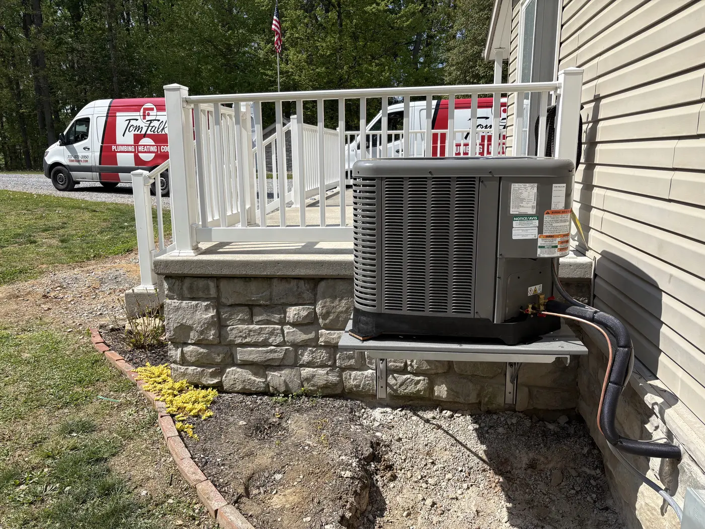Two branded Tom Falk service vans and an American flag beside a newly installed air-conditioning condenser.